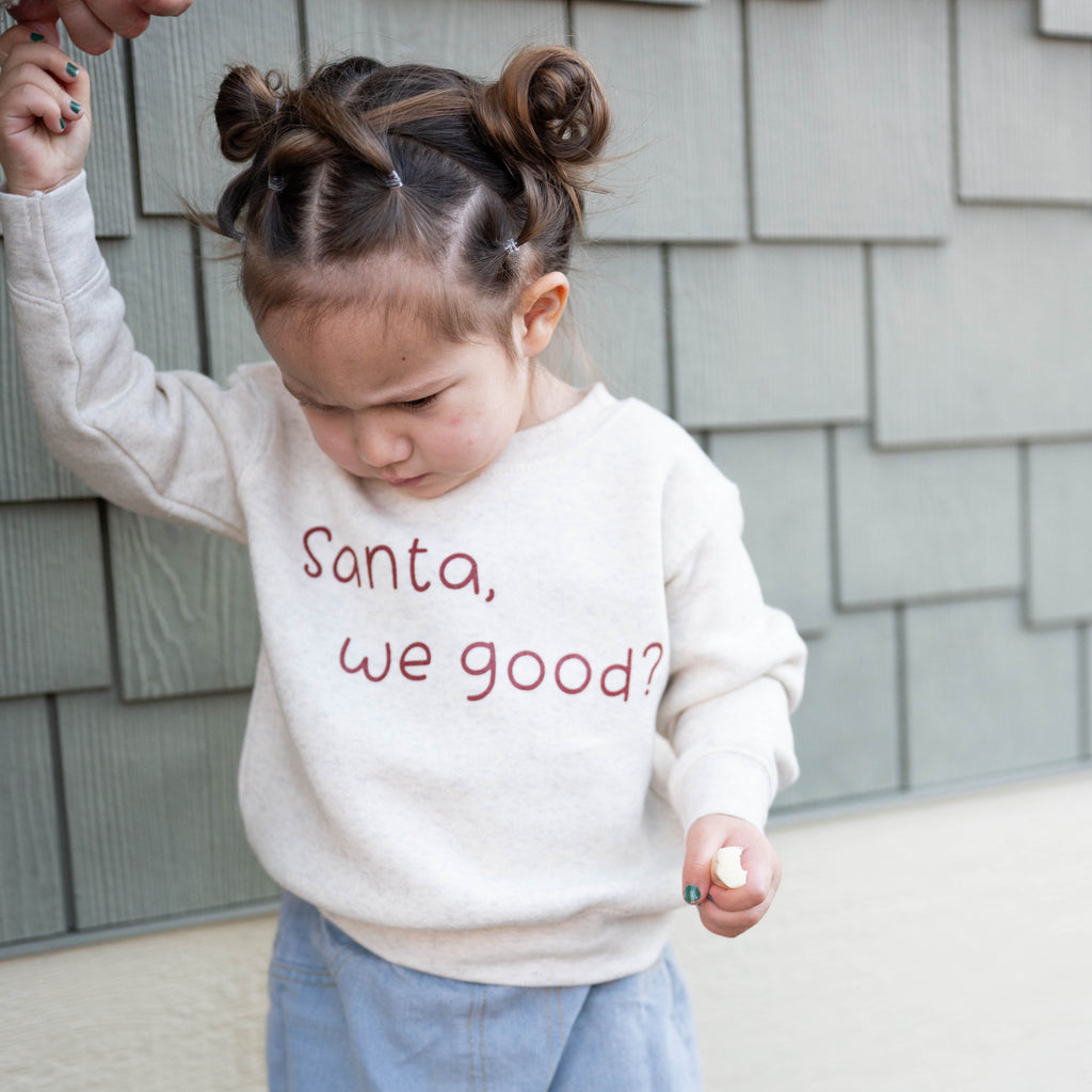 Child wearing a white sweatshirt with 'Santa, we good?' text, standing against a gray wall.
