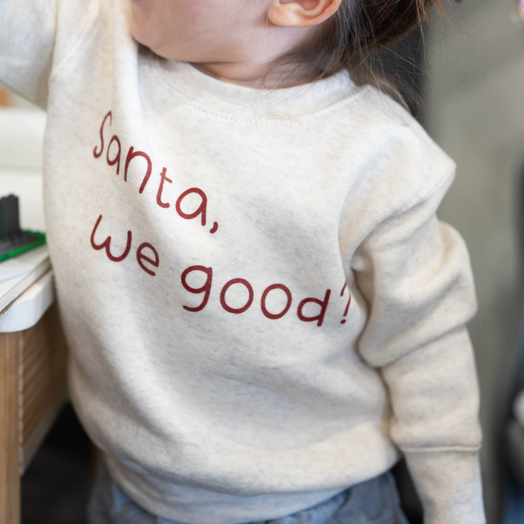 Child wearing a sweater with 'Santa, we good?' text in a casual setting